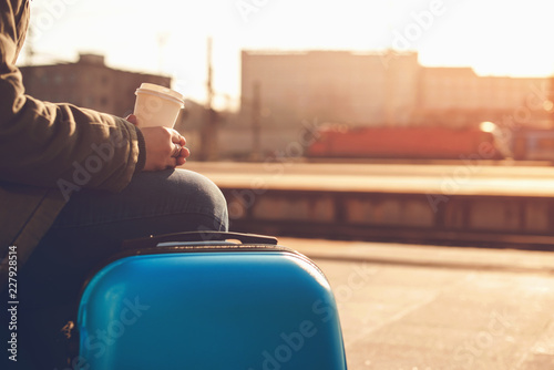 Woman waiting at railway station. Travel by train. Girl drinks coffee and waiting train. Woman with blue luggage suitcase in morning. Lifestyle, travelling, vacation and journey concept. Copy space.