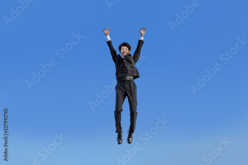 青空をバックにバンザイしながらジャンプするスーツ姿の若いビジネスマン1人。成功・達成・成就・元気・健康イメージ