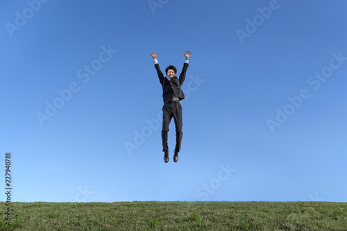 青空をバックにバンザイしながらジャンプするスーツ姿の若いビジネスマン1人。成功・達成・成就・元気・健康イメージ