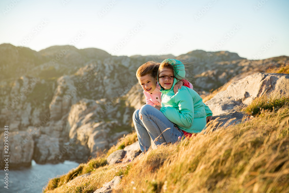 Fototapeta premium Two little girls play on rocky northern seashore. Sit, laugh, hug, explore the coastal rocks. Travel and enjoy a great adventure in Norway. Beautiful view of fjord and mountains in sunset.