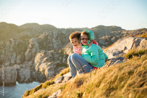 Two little girls play on rocky northern seashore. Sit, laugh, hug, explore the coastal rocks. Travel and enjoy a great adventure in Norway. Beautiful view of fjord and mountains in sunset. © Suzi Media 