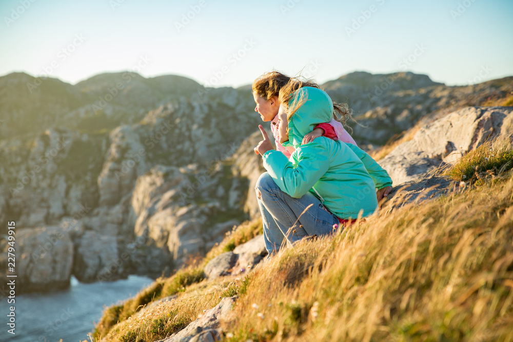 Two little girls play on rocky northern seashore. Sit, laugh, hug, explore the coastal rocks. Travel and enjoy a great adventure in Norway. Beautiful view of fjord and mountains in sunset.