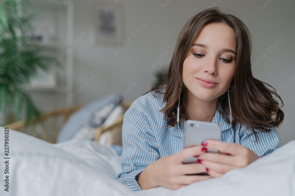 Indoor shot of attractive thoughtful young woman lies in bed, uses modern cell phone, watches video or film online, enjoys good sound, dressed in pyjamas. People, bed time and relaxation concept Indoor shot of attractive thoughtful young woman lies in bed, uses modern cell phone, watches video or film online, enjoys good sound, dressed in pyjamas. People, bed time and relaxation concept