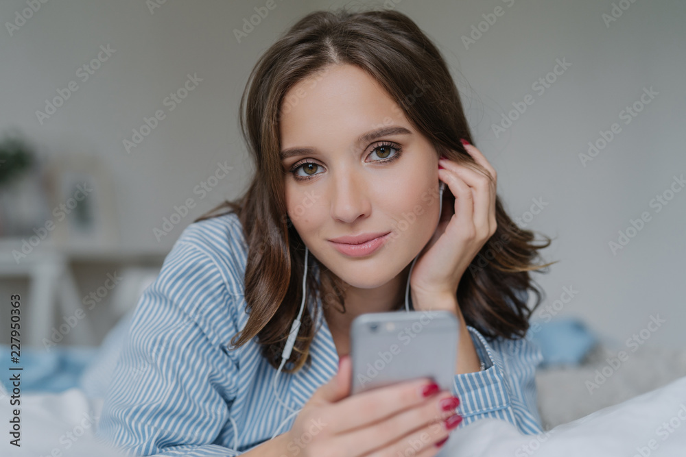 Close up shot of attractive brunette young woman downloads songs from network, uses smart phone for entertainment, dressed in nightclothes, rests in bed, has lazy day, looks seriously at camera Close up shot of attractive brunette young woman downloads songs from network, uses smart phone for entertainment, dressed in nightclothes, rests in bed, has lazy day, looks seriously at camera
