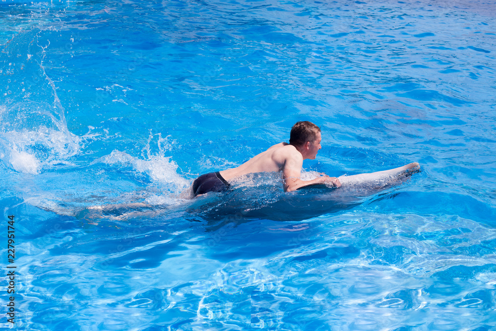 A young man is riding dolphin on the back, boy swimming with dolphin in blue water in water pool, sea, ocean, dolphin saves a man