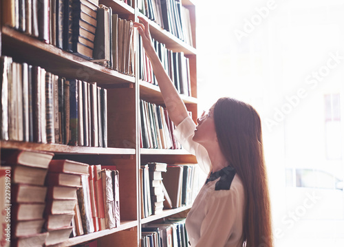 A young student girl is looking for the right book on the shelves of the old university library