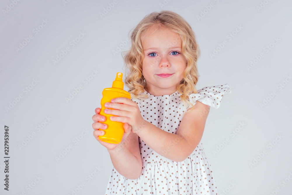 little and beautiful blonde curly hairstyle girl in cute dress holding a bottle sunscreen in hand. blue eyes, perfect skin, on the nose blot spf cream close up portrait on white background in studio little and beautiful blonde curly hairstyle girl in cute dress holding a bottle sunscreen in hand. blue eyes, perfect skin, on the nose blot spf cream close up portrait on white background in studio