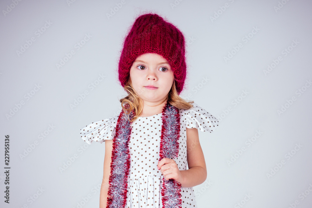 Beautiful little girl in dress and red knitted hat in studio on white background. Happy little smiling female child with shiny Christmas tinsel. Christmas miracles Happy New Year