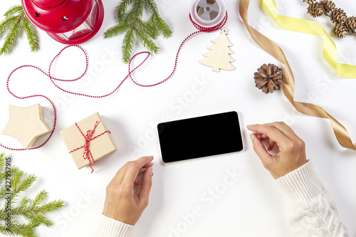 Christmas planing, online shopping concept. Woman hands with mobile phone, christmas presents, decorations on white background