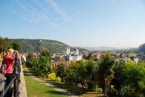 A caucasian woman from backside looking at Sighisoara city panorama with Holy Trinity Orthodox Church and Carpathian Mountains, Romania