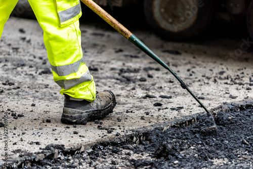 Closeup of construction worker with neon yellow green pants clothing, boots, raking hot black wet tar and asphalt, bitumen in city London