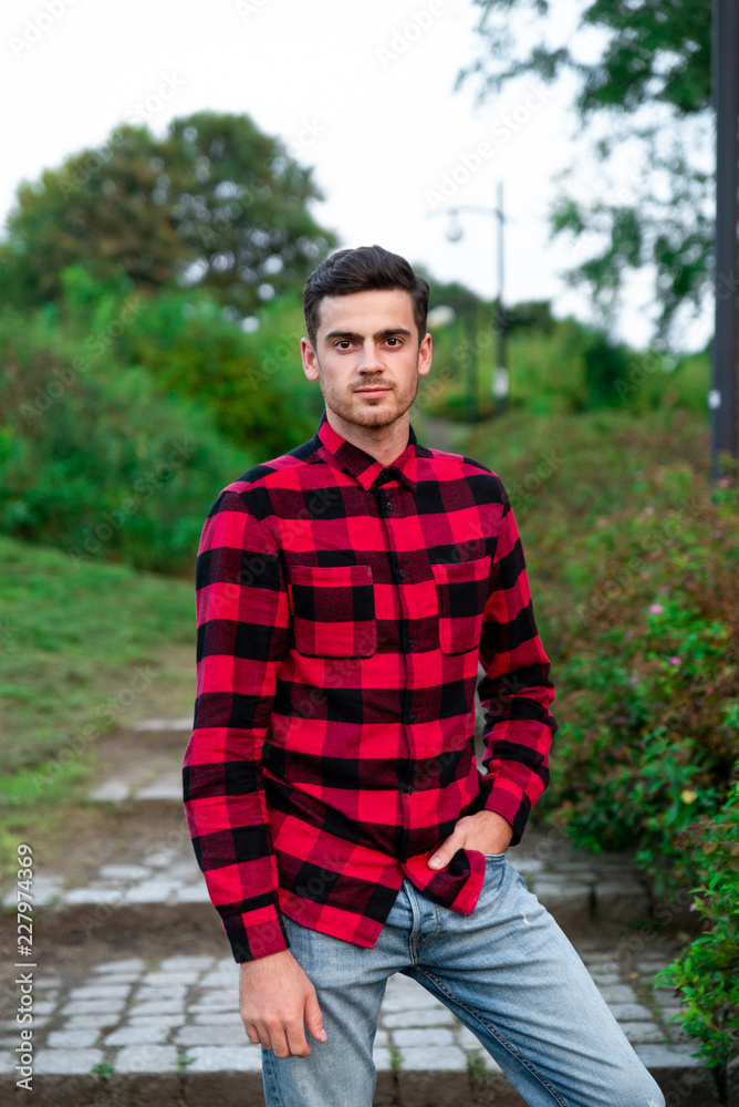Portrait of stylish male student with bread posing outdoors, looking at camera. Handsome man wearing in red checked shirt walking in autumn park. Concept of study, and mans fashion. Portrait of stylish male student with bread posing outdoors, looking at camera. Handsome man wearing in red checked shirt walking in autumn park. Concept of study, and mans fashion.