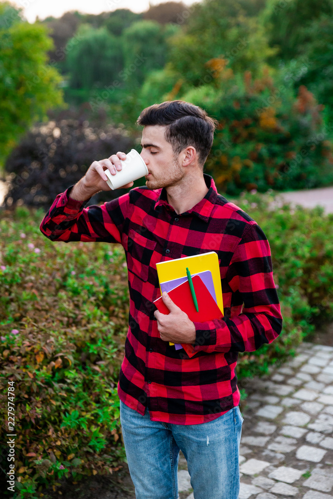 Fototapeta premium Happy student drinking hot coffee and resting after studying. Handsome and stylish brunet male wearing in red checked shirt walking in autumn park with notes and showing at camera cup of coffee. 