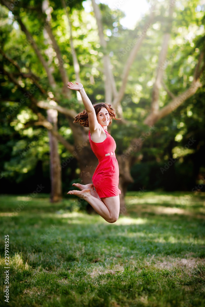 Happy girl jumping for joy and raising her arms. In the background the trees and the green woods of the park. Girl with freckles and red hair, wearing a short red dress. Happy girl jumping for joy and raising her arms. In the background the trees and the green woods of the park. Girl with freckles and red hair, wearing a short red dress.