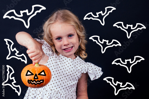 Cute cheerful little witch little girl blonde with blue eyes angel holding a pumpkin with scary grimassy on black background bats flittermouse behind in studio shot.halloween school party photo