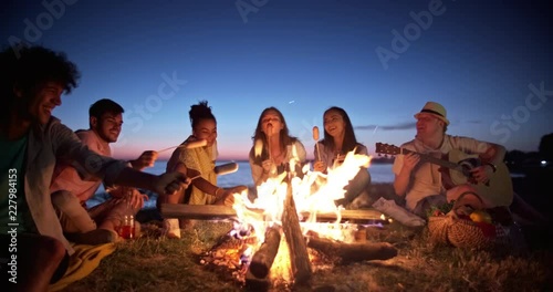 Group of six old multi-ethnic friends sitting near bonfire, roasting sausages, having a summer beach party, talking, enjoying their time 4k