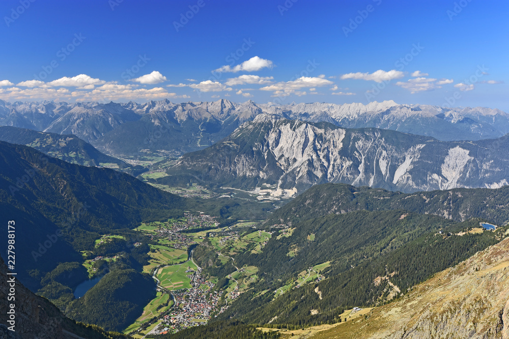 View from the summit of Acherkogel mountain (Tyrol, Austria) at a beautiful day to Inn and Oetztal valley with the village of Oetz. Colorful alpine landscape with rocky mountains, pastures, blue sky.
