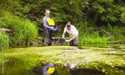 Man and woman scientist environmentalist standing in a river. Woman taking sample of water and pouring it into the test tube. Man holding toolbox