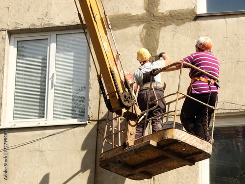 Women plasterers in orange helmets and safety harness with a putty knife and paint roller on a crane putty the seams of a facade old panel  house in the summer day against the windows bottom vie photo