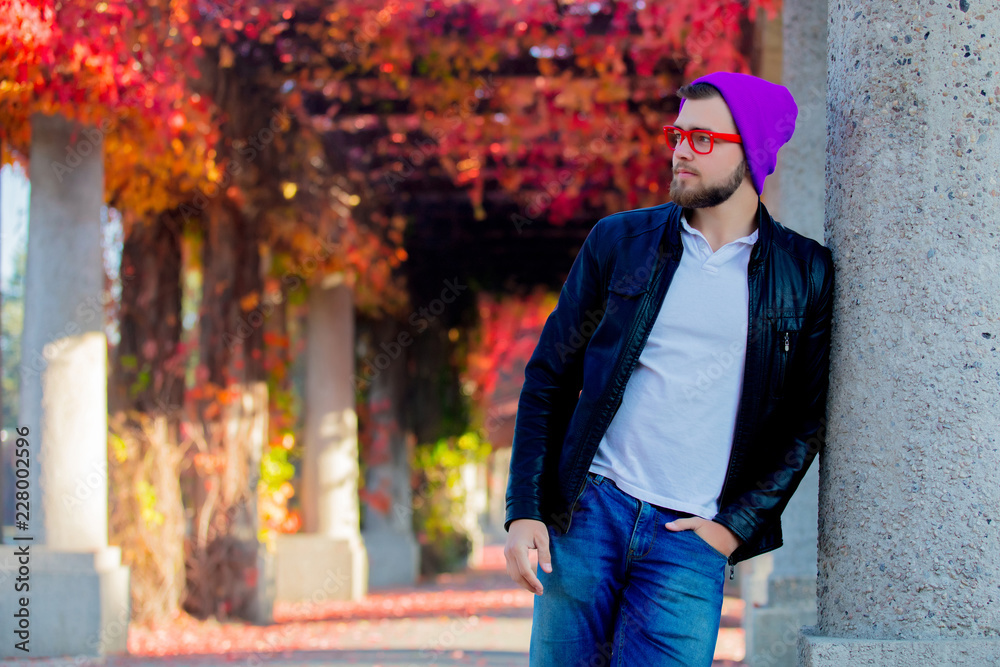 Obraz premium Young white guy in glasses and hat in a park with yellow trees on background. Autumn season time