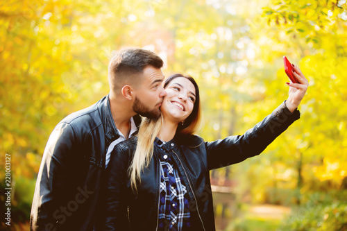 Young white couple make a selfie on mobile phone in a park with yellow trees on background. Autumn season time