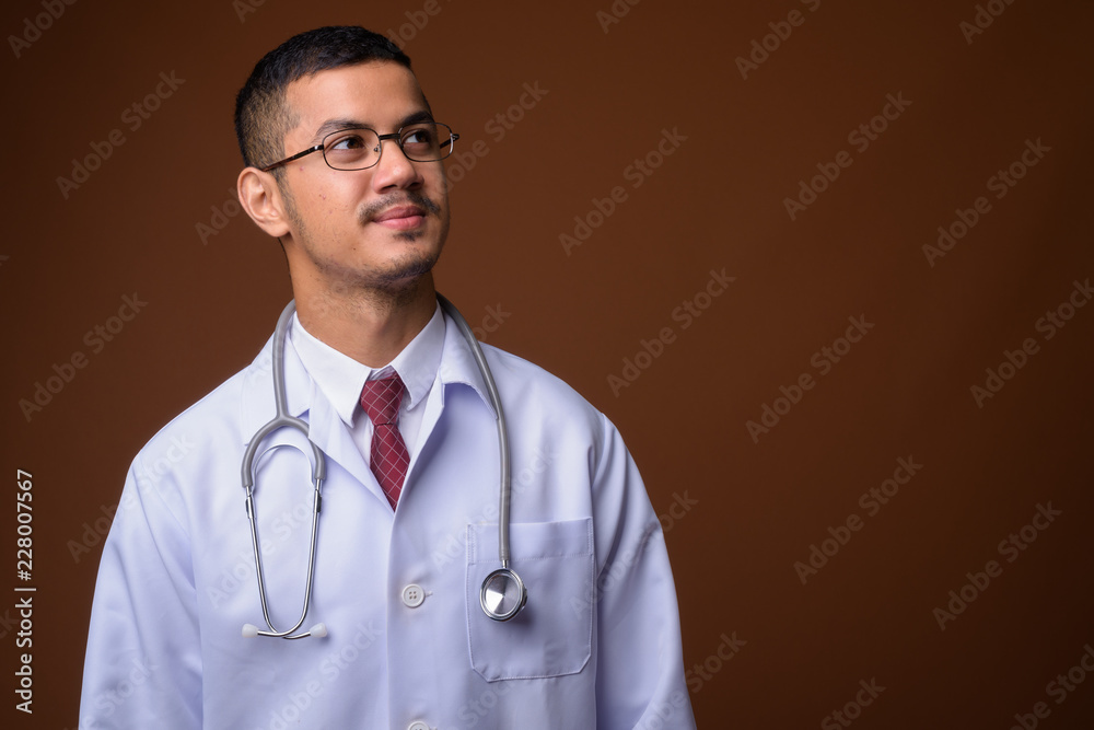 Young multi-ethnic Asian man doctor against brown background Young multi-ethnic Asian man doctor against brown background