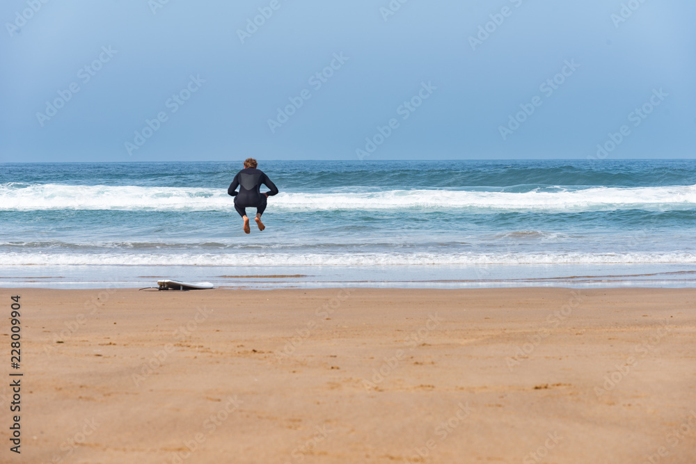 surfer jumping on the beach next to his board with the sea in the background