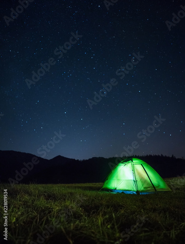 Camping tent with light inside is on the mountain valley under night starry sky