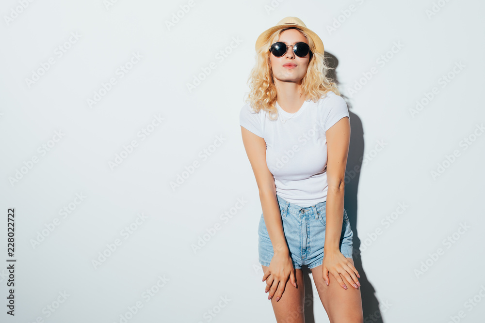 Portrait of a smiling attractive woman in sunglasses, summer clothes and hat posing while standing isolated over white background Portrait of a smiling attractive woman in sunglasses, summer clothes and hat posing while standing isolated over white background