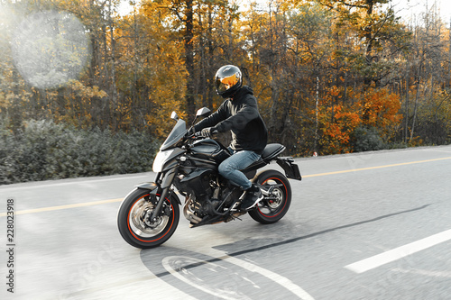 Driver riding motorcycle on empty road in beautiful autumn forest.