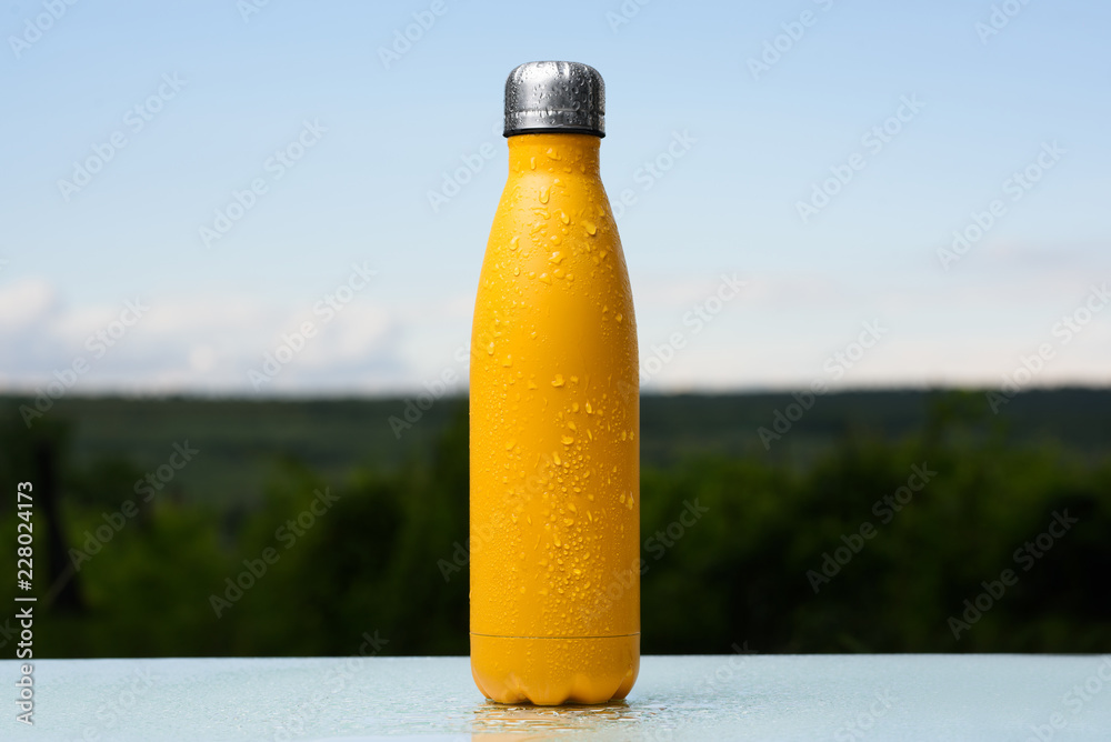 Thermo stainless bottle, sprayed with water. Sky and forest on background. On the glass desk. Thermos of matte yellow color. Thermo stainless bottle, sprayed with water. Sky and forest on background. On the glass desk. Thermos of matte yellow color.