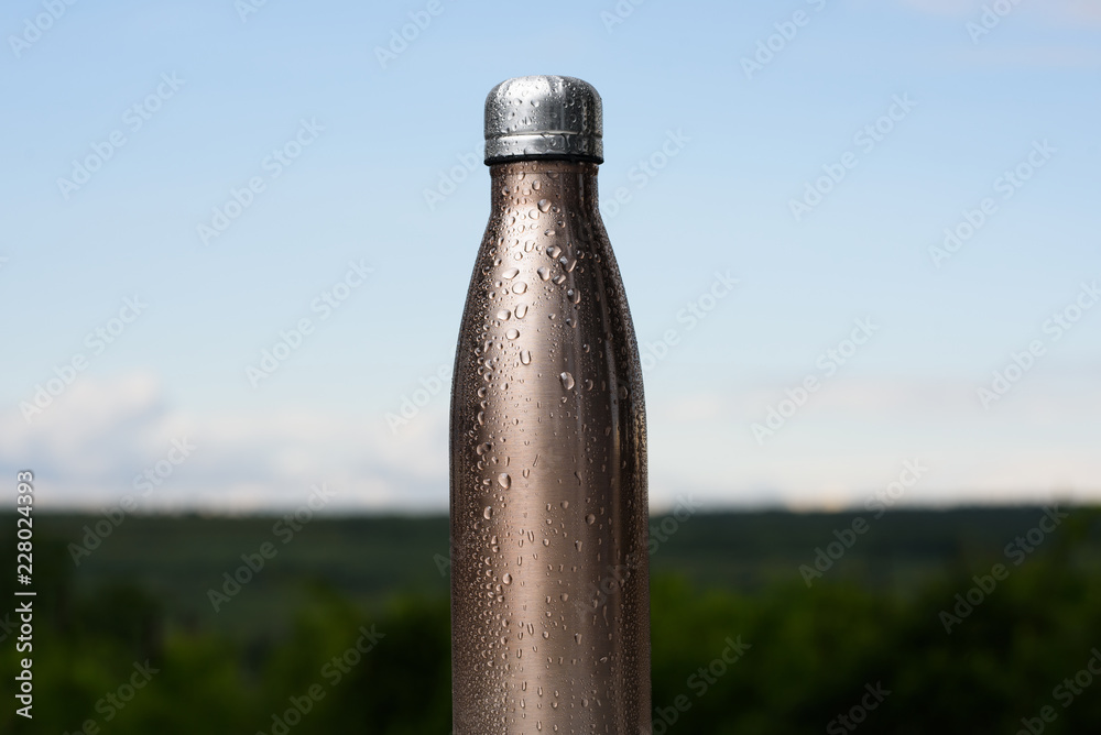 Thermo stainless bottle, sprayed with water. Sky and forest on background. Thermos of maro color. Thermo stainless bottle, sprayed with water. Sky and forest on background. Thermos of maro color.