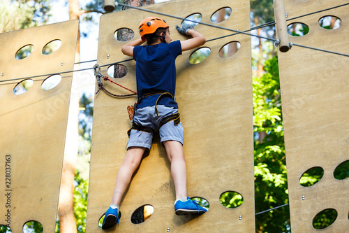 Child in forest adventure park. Kid in orange helmet and blue t shirt climbs on high rope trail. Agility skills and climbing outdoor amusement center for children. young boy plays outdoors.