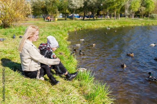 The mother with daughter sitting on grass and looking at lake with ducks
