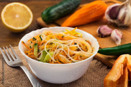 Appetizer of rice noodles in white bowl, fresh vegetables on wooden background.