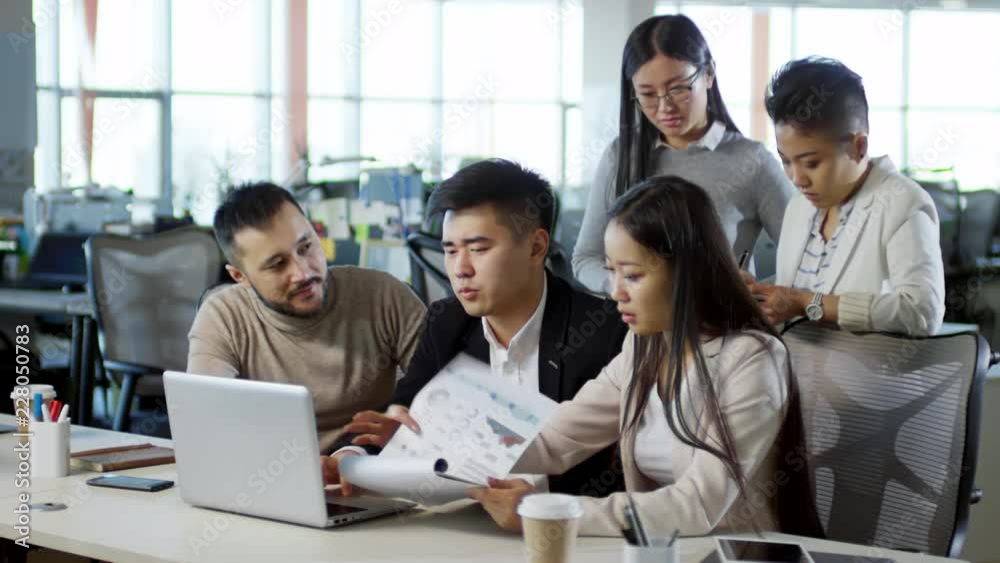 Five Asian business people discussing project and analyzing work results together at desk in office, medium shot