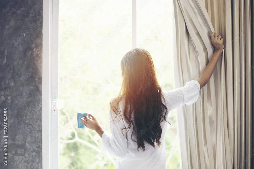 Asian women drinking coffee and wake up in her bed fully rested and open the curtains in the morning to get fresh air on sunshine - obrazy, fototapety, plakaty 