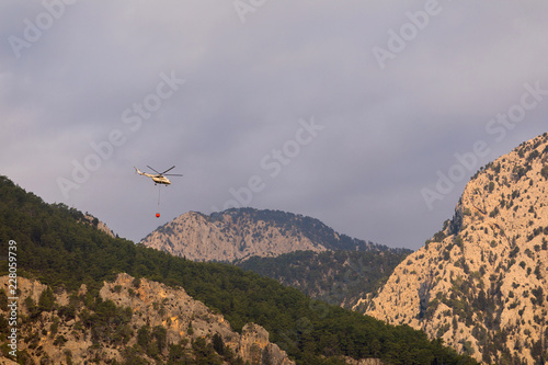 A fire helicopter with a full basket of water flies to extinguish a forest fire against the backdrop of mountains, painted in bright orange sun color. Rescue operation. Forest Fire Prevention