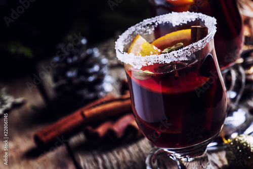 Christmas and New Year festive drink, hot mulled wine with cinnamon, cardamom, orange and anise star on background of green spruce branches and lights, rustic wooden table, selective focus