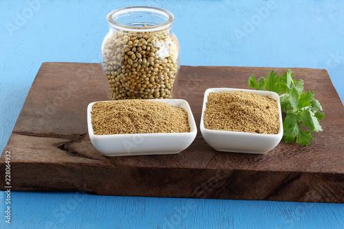 Coriander powder in two ceramic bowls and coriander leaves on a wooden table and seeds in a bottle.