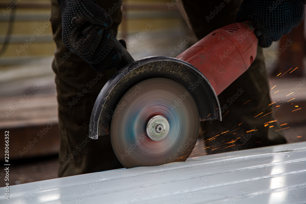 A young male builder in working clothes and black gloves grinds a metal product with angle grinder in construction site