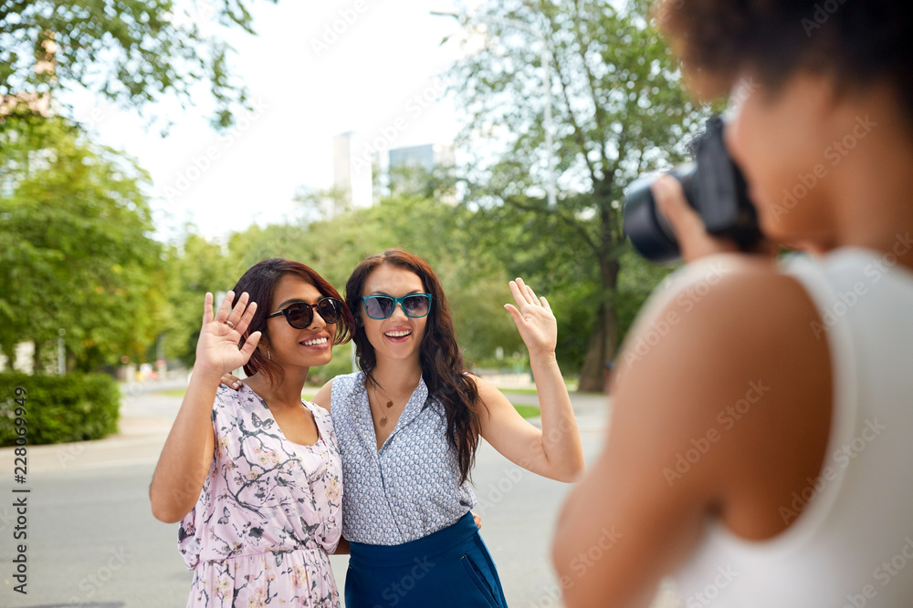 female friendship, technology and people - woman with camera photographing her friends waving hands in summer park