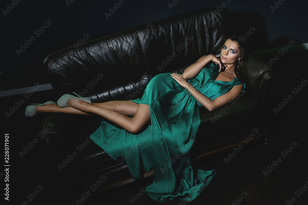 Naklejka premium Woman wearing beautiful green silk dress is posing on leather couch