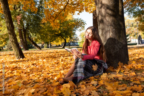 Beautiful young brunette girl sitting on fallen autumn leaves in park  reading book.