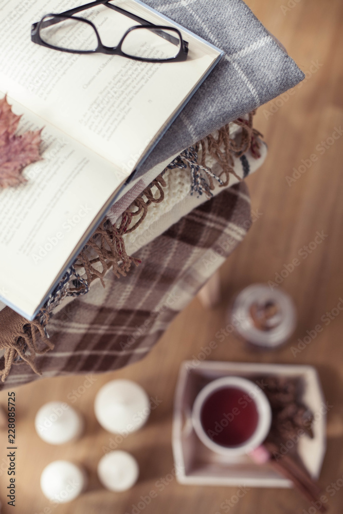 Plaids. A cup of hot tea. Autumn cozy interior. On a wooden chair is a stack of warm blankets. Candles, leaves, cones, basket, cinnamon. Book and glasses. Autumn. Winter.