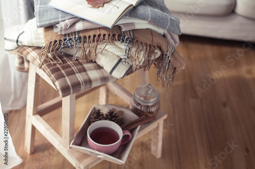 Plaids. A cup of hot tea. Autumn cozy interior. On a wooden chair is a stack of warm blankets. Candles, leaves, cones, basket, cinnamon. Book and glasses. Autumn. Winter.