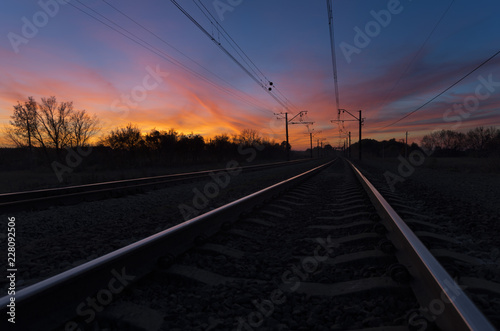 Dark silhouettes railway infrastructure against background of dramatic sunset. View of railroad going straight away to sun and beyond the horizon. The colorful sky with clouds at sundown