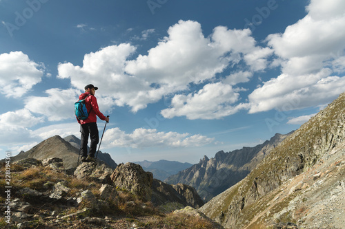 A bearded man in sunglasses and a cap with a backpack stands on top of a rock and looks into a rocky valley high in the mountains. The concept of tourism and easy trekking in the mountains outdoor