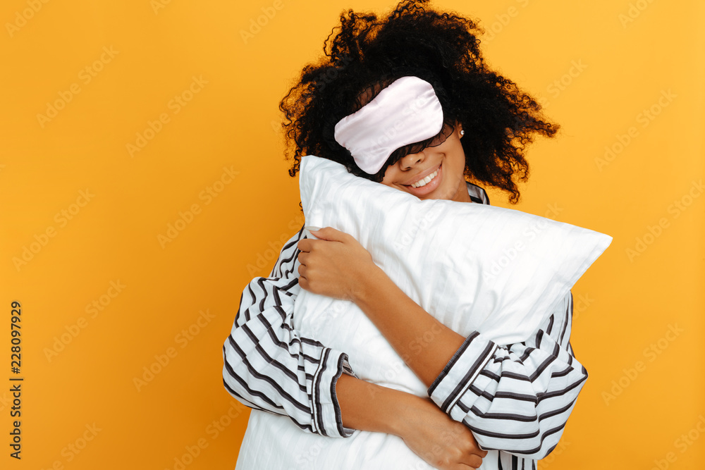 Sleeping. Dreams. Woman portrait. Afro American girl in pajama and sleep mask is hugging a pillow and smiling, on a yellow background - obrazy, fototapety, plakaty 