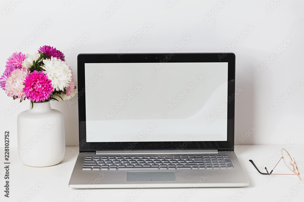 Fototapeta premium Feminine workplace with laptop with white empty screen and flowers in the vase